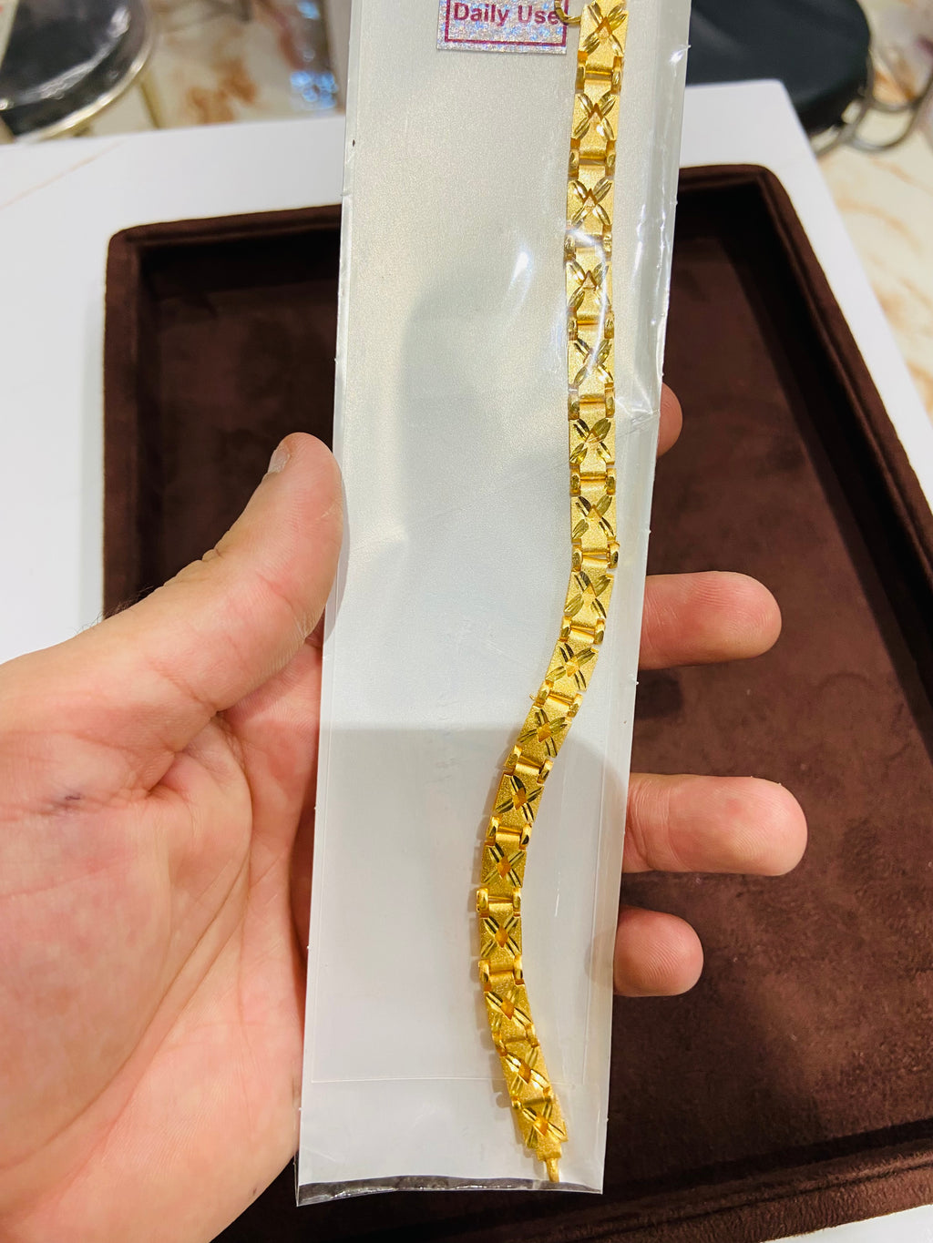 Gold bracelet in a clear plastic packaging held by a hand on a brown surface.