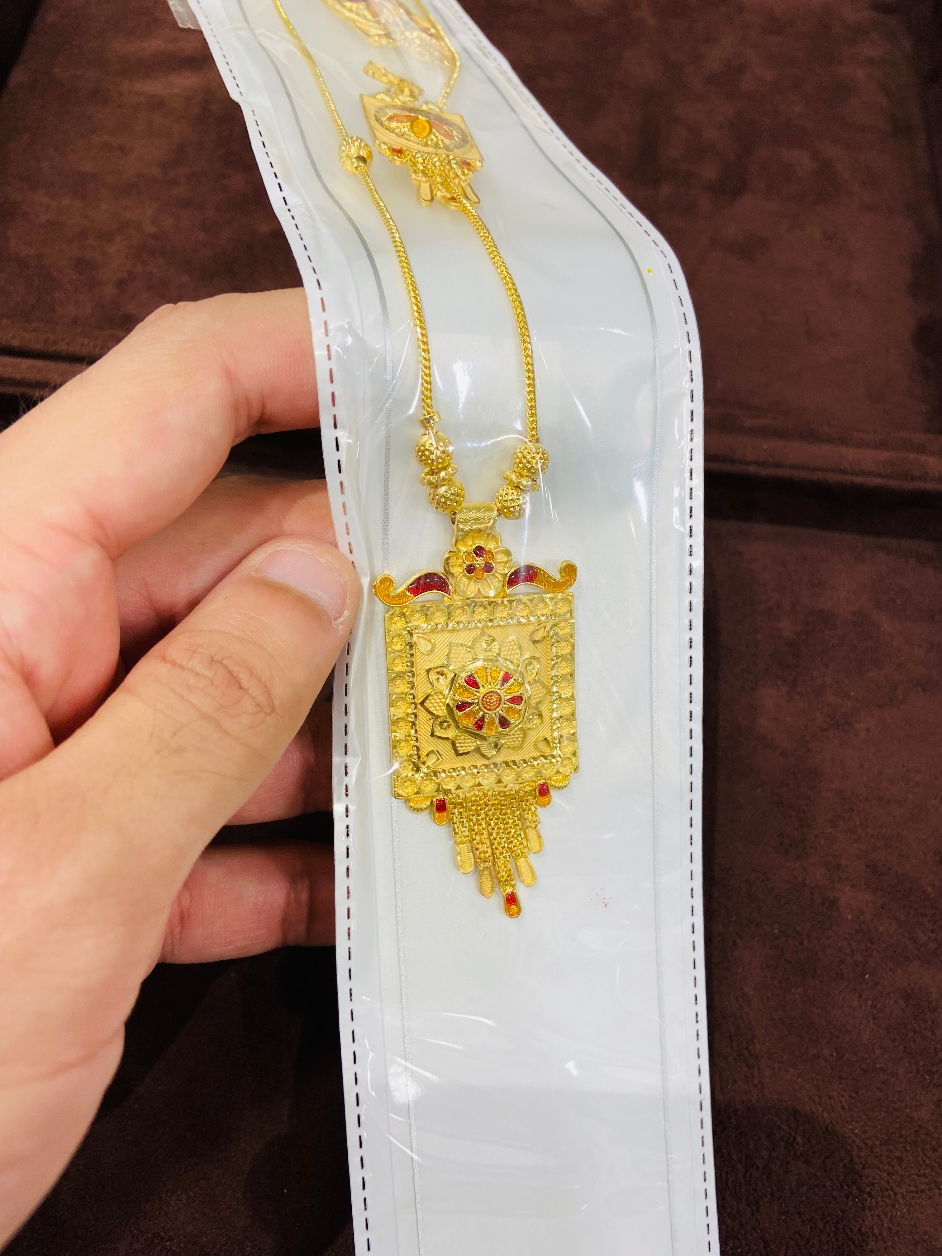Gold necklace with intricate design held by a hand on a white card against a brown background