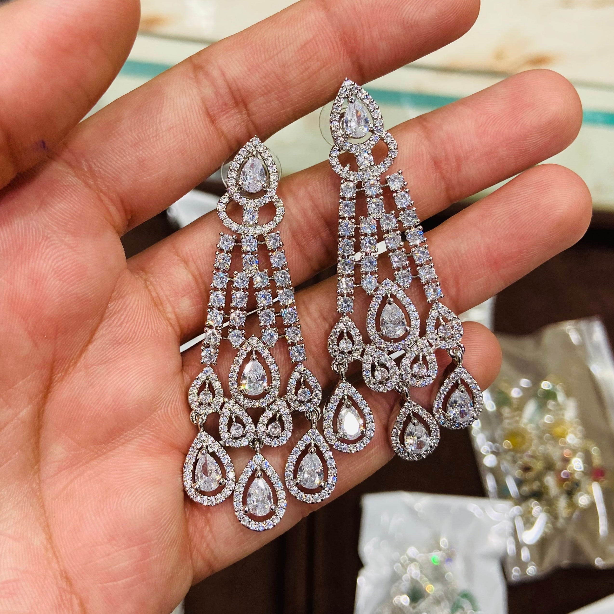 Delicate silver earrings with clear stones held in a hand against a blurred jewelry store background.
