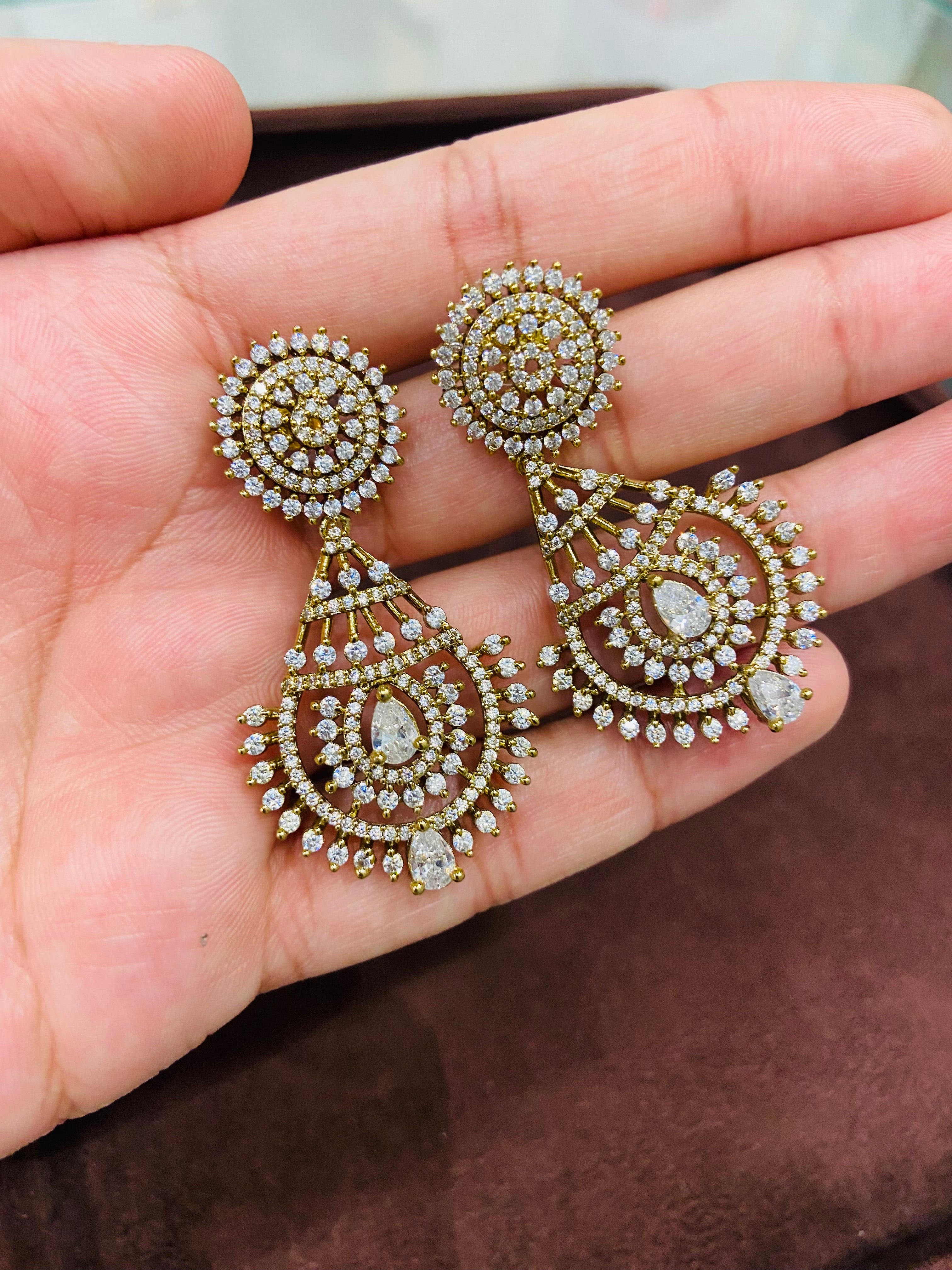Ornate gold and diamond earrings held in a hand against a brown background